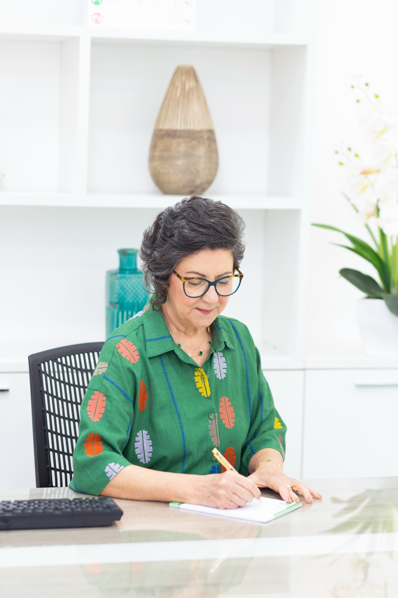 Dra. Sandra Maria Ruiz Prado, pediatra, em retrato profissional usando blusa verde, escrevendo em sua mesa de trabalho